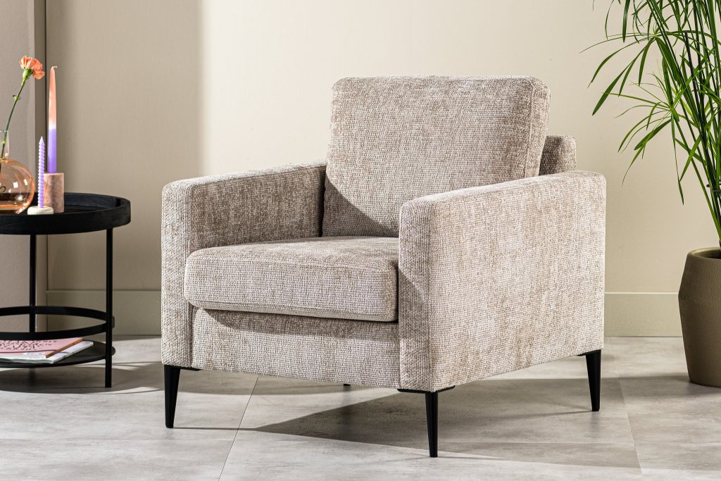 Een beige fauteuil met een structuurstof en zwarte poten staat op een betegelde vloer. Ernaast staat een ronde zwarte tafel met een enkele bloem in een vaas. Een potplant staat ernaast, tegen een lichtgekleurde muur.