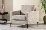 Een beige fauteuil met een structuurstof en zwarte poten staat op een betegelde vloer. Ernaast staat een ronde zwarte tafel met een enkele bloem in een vaas. Een potplant staat ernaast, tegen een lichtgekleurde muur.