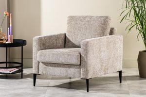 Een beige fauteuil met een structuurstof en zwarte poten staat op een betegelde vloer. Ernaast staat een ronde zwarte tafel met een enkele bloem in een vaas. Een potplant staat ernaast, tegen een lichtgekleurde muur.