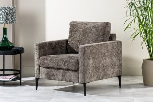 Een grijze gestoffeerde fauteuil met slanke zwarte poten staat op een betegelde vloer. Links ervan staat een ronde zwarte bijzettafel met een groene glazen vaas. Een hoge potplant rechts voegt een vleugje groen toe.