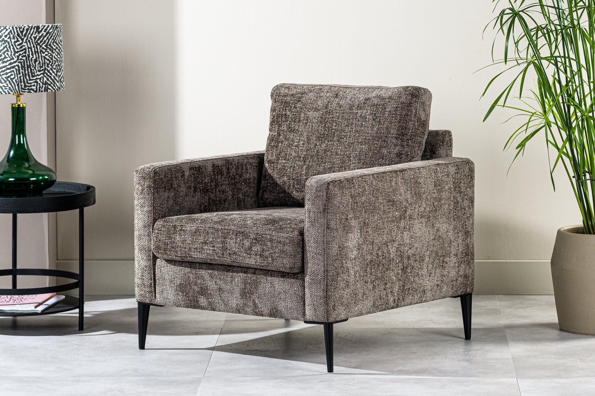 Een grijze gestoffeerde fauteuil met slanke zwarte poten staat op een betegelde vloer. Links ervan staat een ronde zwarte bijzettafel met een groene glazen vaas. Een hoge potplant rechts voegt een vleugje groen toe.