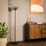 Een moderne woonkamer met een strakke zwarte staande lamp, een houten dressoir en een potplant. De muur is beige met een geometrisch kunstwerk. Schaduwen met lamellen werpen zich over de scène.