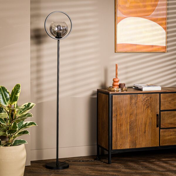Een moderne woonkamer met een strakke zwarte staande lamp, een houten dressoir en een potplant. De muur is beige met een geometrisch kunstwerk. Schaduwen met lamellen werpen zich over de scène.