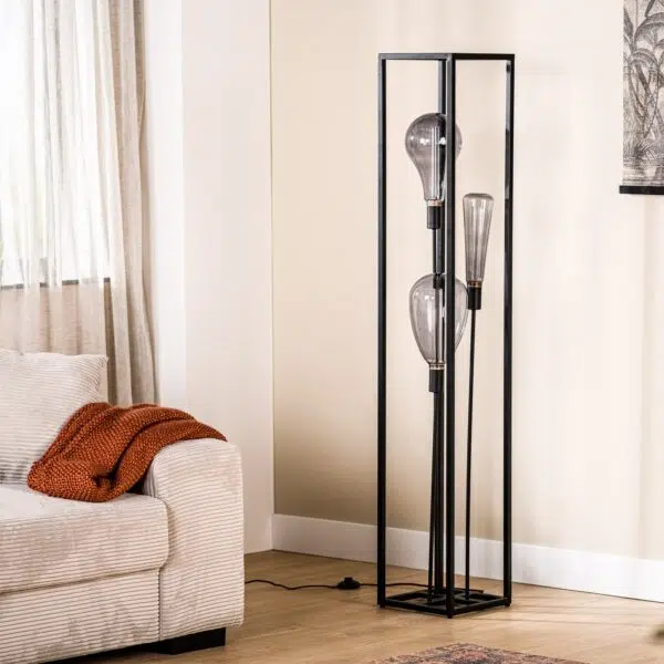 Een moderne staande lamp met drie grote lampen in een minimalistisch zwart frame staat in een lichte woonkamer. Ernaast staat een beige bank met witte en roestkleurige kussens. Licht filtert door een raam met doorschijnende gordijnen. Er hangt een monochrome wandkunst boven.
