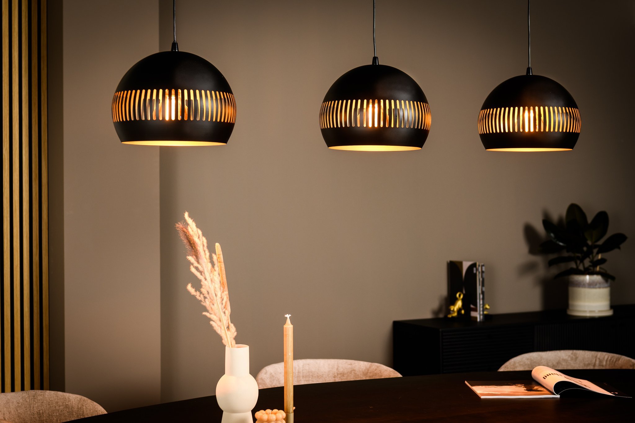 Drie zwarte hanglampen met gouden details aan de binnenkant hangen boven een donkere houten tafel. Op de tafel staat een witte vaas met gedroogde planten, een kaars en een tijdschrift. Op de achtergrond staat een dressoir en een plant op een plank.