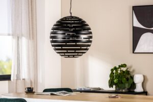 Moderne eethoek met een ronde, zwarte, lamellen hanglamp boven een houten tafel. Op de tafel staan een tijdschrift, een kleine plant en een kopje. Op de achtergrond zijn een witte buste en abstracte kunst zichtbaar op een beige muur.