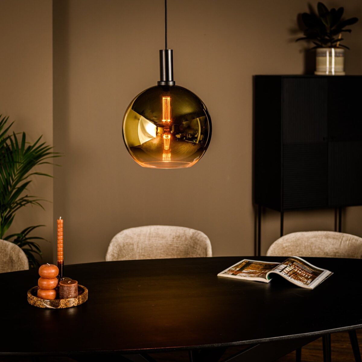 Een gezellige eetkamer met een ronde zwarte tafel, een moderne amberkleurige glazen hanglamp erboven en een kleine stapel kaarsen op tafel. Een tijdschrift ligt open naast de kaarsen. Beige stoelen omringen de tafel en planten versieren de kamer.