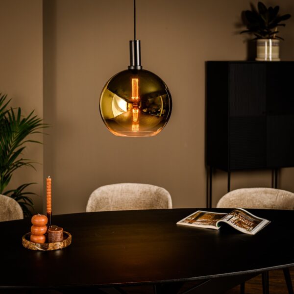 Een moderne eetkamer heeft een donkere ovale tafel met beige stoelen. Een gouden, bolvormige hanglamp hangt erboven. Een brandende kaars staat op de tafel naast een open boek. Een plant en een kast staan op de achtergrond tegen een beige muur.