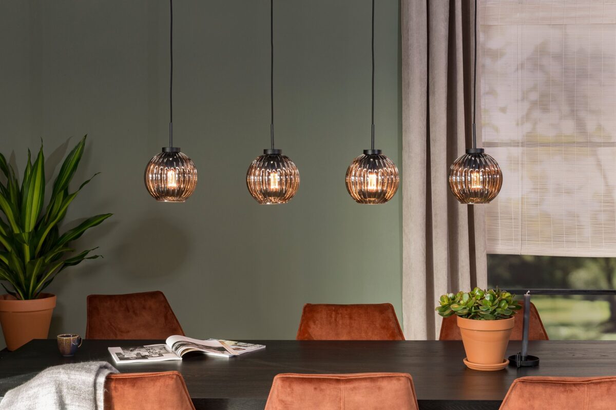 Een eetkamer met een donkere tafel omringd door bruine stoelen. Vier bolvormige hanglampen hangen boven de tafel. Een potplant staat op de tafel en een andere staat op de grond. Een tijdschrift ligt open op de tafel en op de achtergrond is een raam.