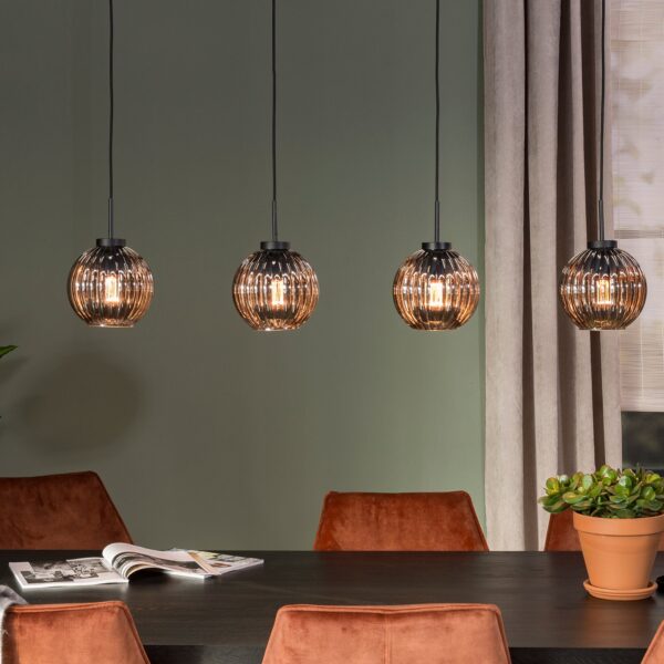 Een eetkamer met een donkere tafel omringd door bruine stoelen. Vier bolvormige hanglampen hangen boven de tafel. Een potplant staat op de tafel en een andere staat op de grond. Een tijdschrift ligt open op de tafel en op de achtergrond is een raam.