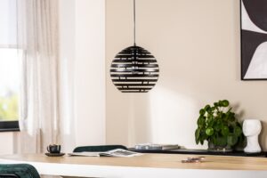 Een moderne eetkamer heeft een zwart-wit gestreepte bolvormige hanglamp boven een lichte houten tafel. De tafel bevat een kopje, een tijdschrift en onderzetters. Een potplant en abstracte kunst staan op de achtergrond. Natuurlijk licht filtert door een raam.