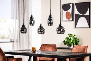 Moderne eetkamer met een donkere houten tafel, vier bruine gestoffeerde stoelen en vijf hangende hanglampen. Er staat een potplant op tafel. Abstracte kunst hangt aan de crèmekleurige muren en transparante gordijnen bedekken een raam.