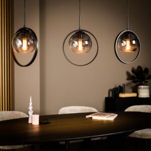 Een moderne eethoek met een donkere houten tafel en drie hanglampen met ronde frames en zichtbare gloeilampen. Op de tafel staan een decoratieve witte kaars, een boek en een klein roze beeldje. Een zwarte kast bevat een plant op de achtergrond.