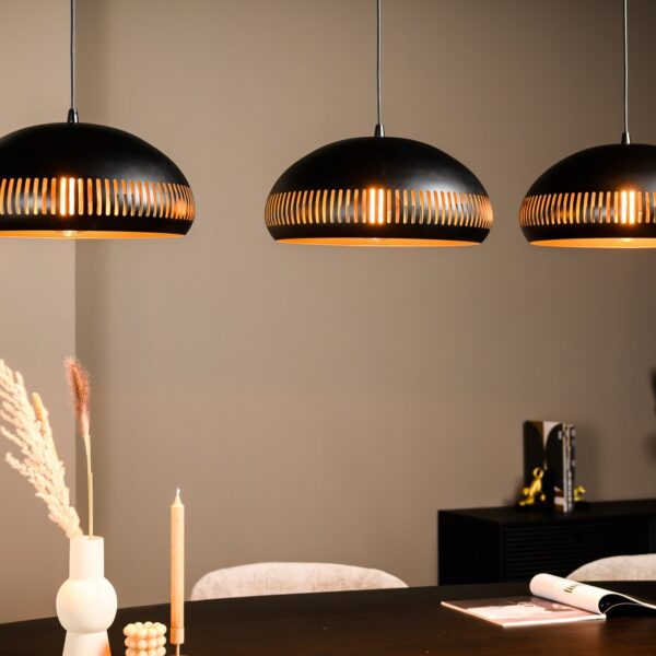 Een eetkamer heeft drie hanglampen met zwarte en gouden accenten die boven een donkere tafel hangen. Op de tafel liggen gedroogde grassen in een vaas, een beige kaars, decoratieve kralen en een open tijdschrift. De achtergrond is neutraal met subtiele decoratie.