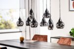 Een moderne eethoek met een zwarte tafel, geaccentueerd door bruine fluwelen stoelen. Erboven zorgen verschillende hangende glazen hanglampen voor verlichting. Een tijdschrift en een gele mok staan op tafel. Een raam en een plant zorgen voor natuurlijk licht en groen.