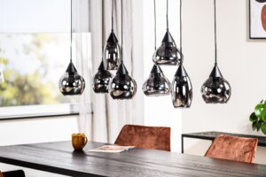 Een moderne eethoek heeft een zwarte houten tafel met een bruine mok en een tijdschrift. Erboven hangen verschillende zwarte hanglampen met glazen kappen. Op de achtergrond staan bruine stoelen, een raam met doorzichtige gordijnen en een plant.