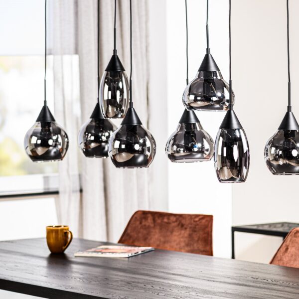 Een moderne eethoek met een zwarte tafel, geaccentueerd door bruine fluwelen stoelen. Erboven zorgen verschillende hangende glazen hanglampen voor verlichting. Een tijdschrift en een gele mok staan op tafel. Een raam en een plant zorgen voor natuurlijk licht en groen.