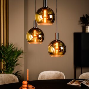 Drie bolvormige hanglampen hangen boven een ronde tafel met een decoratieve kaarsenhouder en een open tijdschrift. De lampen zijn goudkleurig met zichtbare filamenten, wat een warme sfeer creëert. Een potplant en een houten paneel staan op de achtergrond.