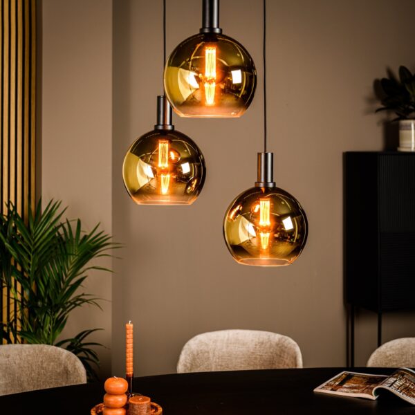 Drie amberkleurige glazen hanglampen met zichtbare filamenten hangen boven een ronde zwarte tafel. Een kleine opstelling met oranje kaarsen staat op de tafel. Beige stoelen omringen de tafel en groene planten accentueren de warm getinte kamer.