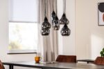 Een moderne eetkamer heeft een cluster van glimmende zwarte hanglampen die boven een donkere houten tafel hangen. Een gouden beker en een tijdschrift liggen op tafel. De kamer heeft een raam met jaloezieën en neutrale gordijnen. Bruine stoelen omringen de tafel.