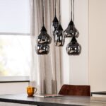 Een moderne eetkamer heeft een cluster van glimmende zwarte hanglampen die boven een donkere houten tafel hangen. Een gouden beker en een tijdschrift liggen op tafel. De kamer heeft een raam met jaloezieën en neutrale gordijnen. Bruine stoelen omringen de tafel.