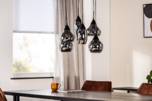 Een moderne eetkamer heeft een cluster van glimmende zwarte hanglampen die boven een donkere houten tafel hangen. Een gouden beker en een tijdschrift liggen op tafel. De kamer heeft een raam met jaloezieën en neutrale gordijnen. Bruine stoelen omringen de tafel.