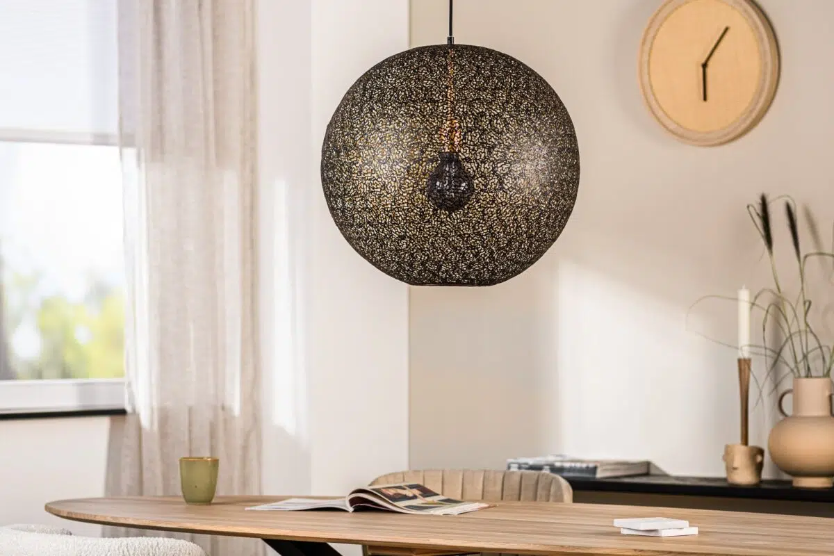 Een gezellige eetkamer met een grote, decoratieve hanglamp boven een houten tafel. De tafel heeft een open tijdschrift en een groene mok. Een ronde wandklok en lichte gordijnen sieren de minimalistische ruimte.