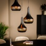 Drie moderne zwarte hanglampen met gleufontwerpen hangen boven een ronde donkere tafel. Zacht warm licht straalt ervan af. Een tijdschrift ligt op de tafel. Groene planten en minimale meubels accentueren de neutrale kamer.