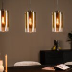 Vier moderne hanglampen met gouden glazen kappen hangen boven een zwarte eettafel. Op de tafel staat een brandende kaars, gedroogde bloemen in een vaas en een open boek. Achter de tafel staat een donker dressoir met decoratie, waaronder een plant en boeken, tegen een beige muur.