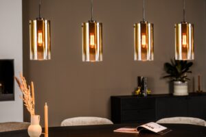 Vier moderne hanglampen met gouden glazen kappen hangen boven een zwarte eettafel. Op de tafel staat een brandende kaars, gedroogde bloemen in een vaas en een open boek. Achter de tafel staat een donker dressoir met decoratie, waaronder een plant en boeken, tegen een beige muur.