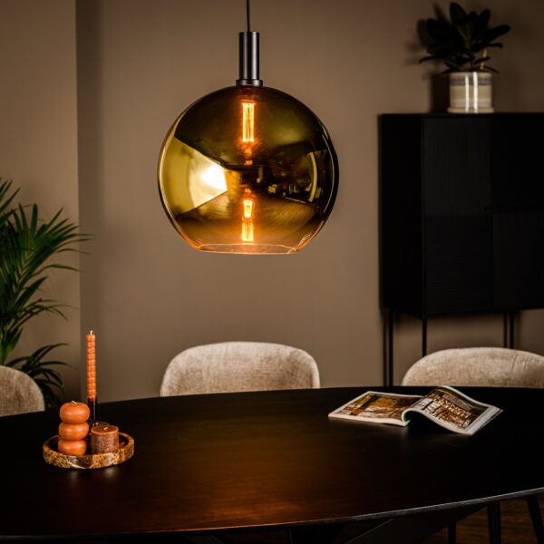 Een moderne eetkamer heeft een grote amberkleurige bolvormige hanglamp boven een donkere ronde tafel. Op de tafel staat een kaarsenhouder met een hoge, onverlichte kaars en een open tijdschrift. Op de achtergrond zijn een donkere kast en een potplant zichtbaar.
