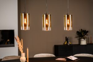 Drie moderne hanglampen met gouden cilindrische kappen hangen boven een donkere tafel. Op de tafel staat een open boek, een vaas met gedroogde planten en een brandende kaars. De achtergrond is een gedempt bruine muur met een kleine plant en decoratie op een zijkast.