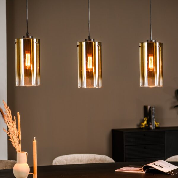 Drie moderne hanglampen met gouden cilindrische kappen hangen boven een donkere tafel. Op de tafel staat een open boek, een vaas met gedroogde planten en een brandende kaars. De achtergrond is een gedempt bruine muur met een kleine plant en decoratie op een zijkast.
