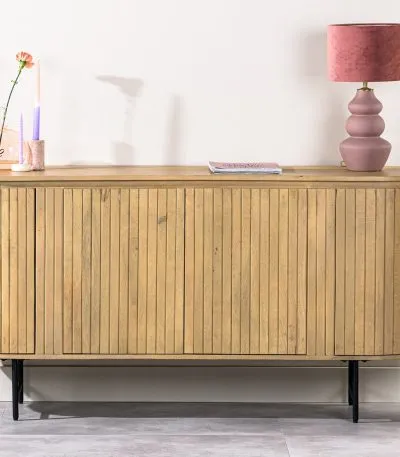 Een houten dressoir met een lattenontwerp staat tegen een lichte muur. Het heeft een roze lamp met een gelaagde basis, een kleine glazen vaas met een bloem en een tijdschrift. Een hoge groene plant staat in een pot aan de rechterkant.