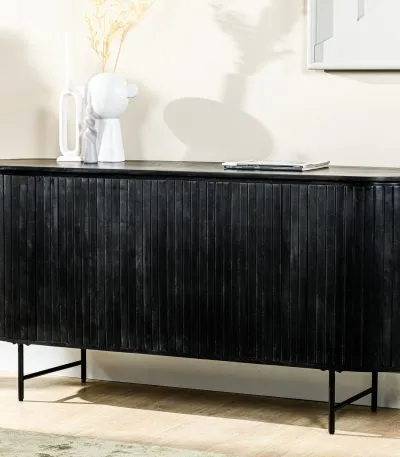 Een strak zwart dressoir met geribbelde textuur staat tegen een beige muur. Het is versierd met een minimalistische witte vaas en decoratieve items erbovenop. Zonlicht filtert door een nabijgelegen raam en werpt zachte schaduwen op de hardhouten vloer.