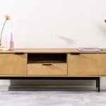 Een minimalistische houten tv-standaard met metalen poten, met twee zijkastjes en een lade in het midden. Versierd met een roze bloem in een vaas, twee hoge kaarsen en een boek. Een potplant staat aan de rechterkant op een betegelde vloer.