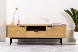 Een minimalistische houten tv-standaard met metalen poten, met twee zijkastjes en een lade in het midden. Versierd met een roze bloem in een vaas, twee hoge kaarsen en een boek. Een potplant staat aan de rechterkant op een betegelde vloer.