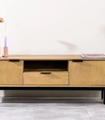 Een minimalistische houten tv-standaard met metalen poten, met twee zijkastjes en een lade in het midden. Versierd met een roze bloem in een vaas, twee hoge kaarsen en een boek. Een potplant staat aan de rechterkant op een betegelde vloer.