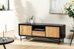Een moderne zwarte mediaconsole met houten lattenkasten en open opbergruimte staat tegen een lichtgekleurde muur. Een kleine decoratieve vaas met groen en een vleugje zonlicht versterken het minimalistische decor.
