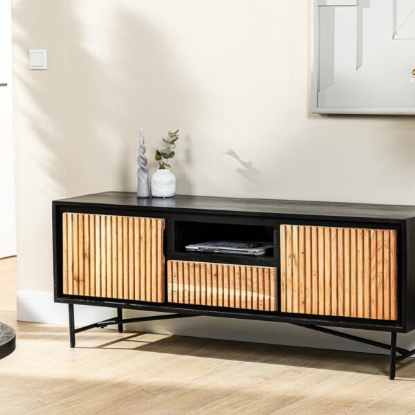 Een moderne zwarte mediaconsole met houten lattenkasten en open opbergruimte staat tegen een lichtgekleurde muur. Een kleine decoratieve vaas met groen en een vleugje zonlicht versterken het minimalistische decor.