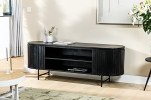 Een moderne, zwarte mediaconsole met een gebogen ontwerp staat tegen een lichtgekleurde muur. Het heeft open planken met een paar tijdschriften en decoratieve items. Een vaas met bloemen staat erbovenop en zonlicht filtert door de gordijnen in de buurt.