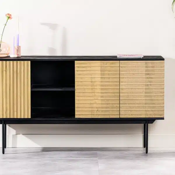 Een modern dressoir met zwarte poten en houten lamellendeuren staat tegen een witte muur. Decoratie-items, waaronder een vaas met een bloem en een kaars, staan erop. Een hoge groene plant in een grijze pot staat rechts.