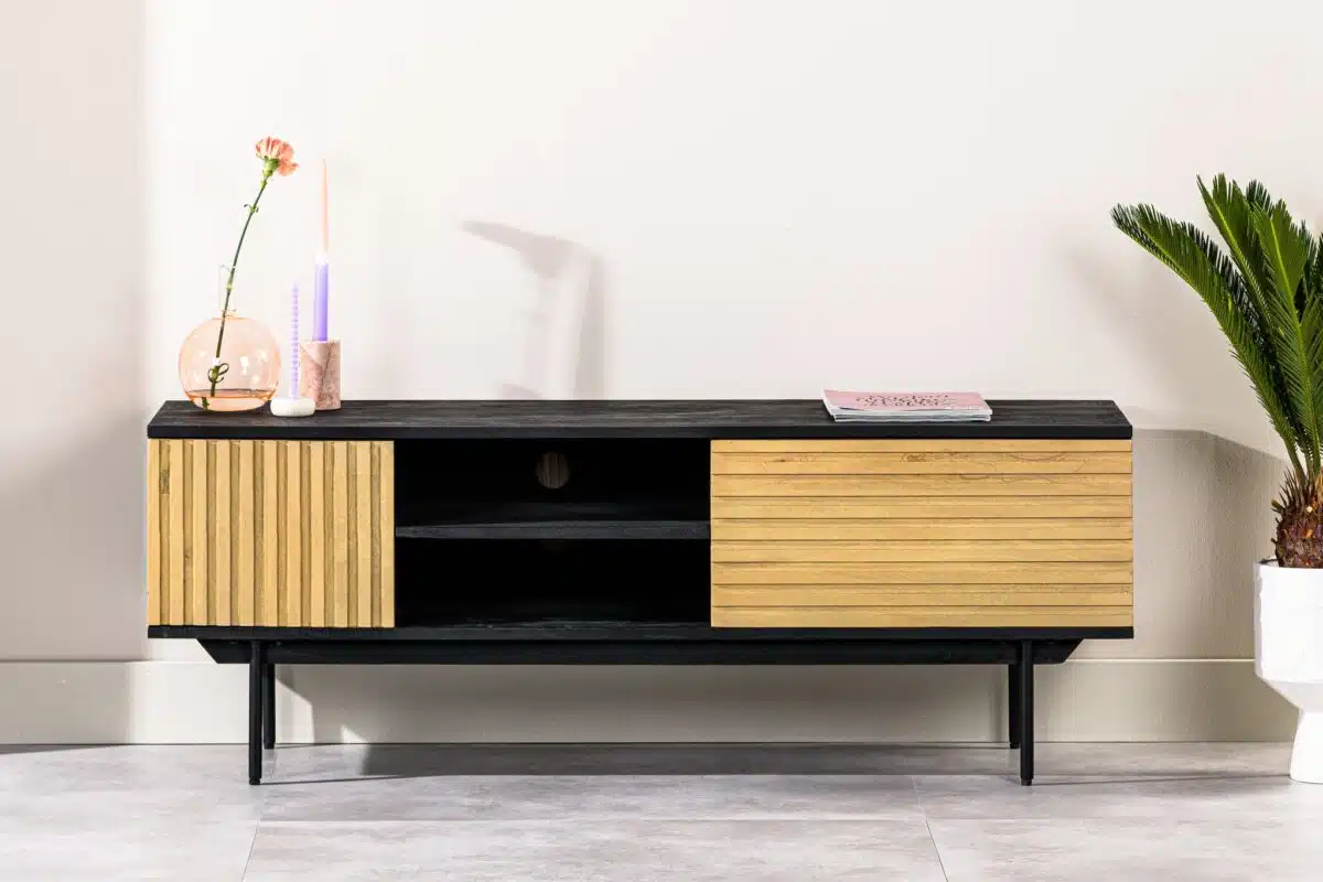 Een moderne zwarte tv-standaard met houten schuifdeuren, met open planken. Bovenop staat een kaars, een kleine roze glazen vaas met een oranje bloem en een boek. Een groene potplant staat ernaast aan de rechterkant. De achtergrond is een lichte muur.