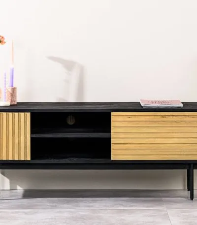 Een moderne zwarte tv-standaard met houten schuifdeuren, met open planken. Bovenop staat een kaars, een kleine roze glazen vaas met een oranje bloem en een boek. Een groene potplant staat ernaast aan de rechterkant. De achtergrond is een lichte muur.