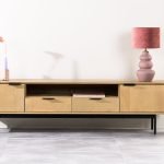 Een moderne houten tv-standaard met twee lades en open planken, met een roze lamp en een vaas met een bloem en een kaars. Het staat tegen een lichte muur, met een grote plant in een pot aan de rechterkant. De vloer is lichtgekleurd.