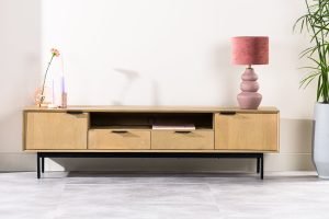 Een moderne houten tv-standaard met twee lades en open planken, met een roze lamp en een vaas met een bloem en een kaars. Het staat tegen een lichte muur, met een grote plant in een pot aan de rechterkant. De vloer is lichtgekleurd.