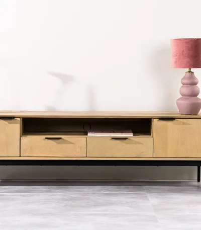 Een moderne houten tv-standaard met twee lades en open planken, met een roze lamp en een vaas met een bloem en een kaars. Het staat tegen een lichte muur, met een grote plant in een pot aan de rechterkant. De vloer is lichtgekleurd.