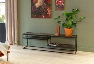 Een moderne woonkamer met een groene muur met twee bloemenschilderijen. Een consoletafel van zwart metaal en hout met een potplant en twee decoratieve kaarsen. Lichte houten vloer en beige gordijnen zijn zichtbaar.