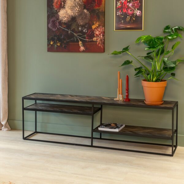 Een moderne woonkamer met een groene muur met twee bloemenschilderijen. Een consoletafel van zwart metaal en hout met een potplant en twee decoratieve kaarsen. Lichte houten vloer en beige gordijnen zijn zichtbaar.