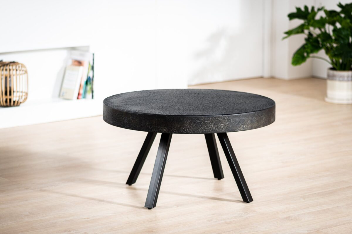 De Salontafel rond 74 cm, Lava, B341 vintage zwart, een ronde zwarte salontafel met vier schuine poten, staat op een lichte houten vloer in een moderne kamer met een witte muur, boeken op een plank en een groene potplant.